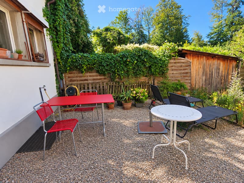 Outdoor patio area with gravel ground, red metal table and chairs, white round table, sun lounger, potted plants, wooden fence, and greenery.