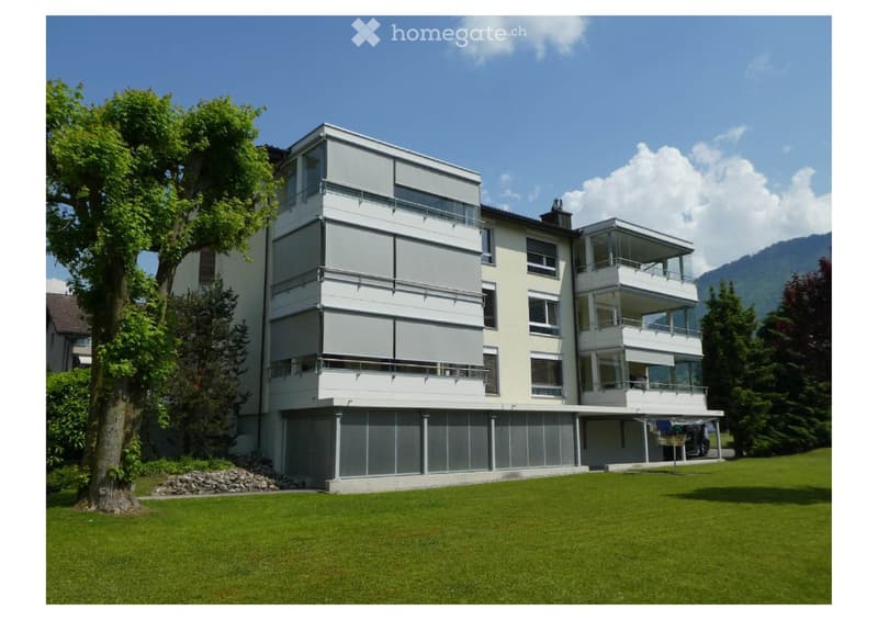 Modern multi-story house with large windows, balconies, and a raised ground floor, surrounded by a well-maintained lawn and trees.