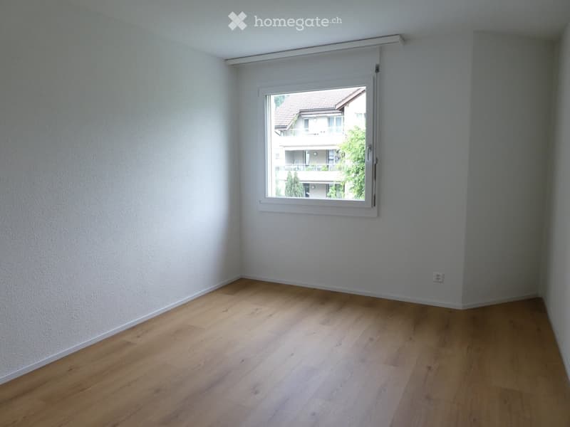 Empty room with light wooden flooring and white walls, featuring a large window with a view of neighboring buildings and greenery.
