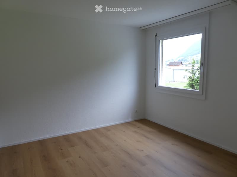 Empty room with light wooden flooring and white walls, featuring a large window with a view of the outdoors.