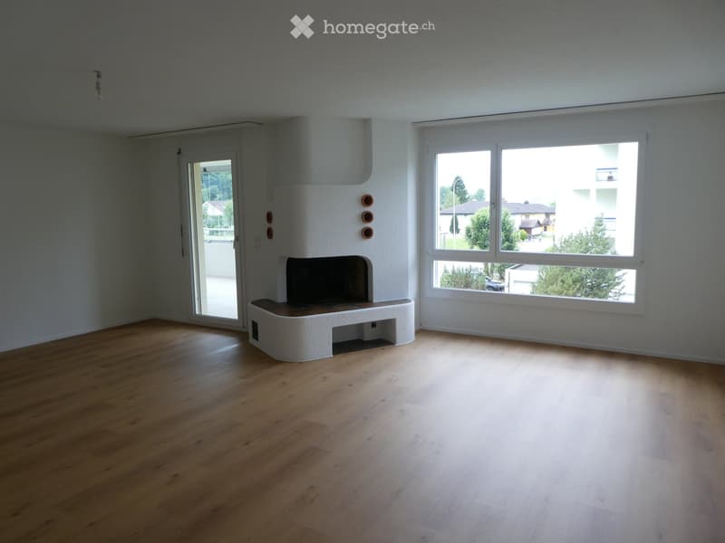Empty room with wooden flooring, white walls, large windows, and a built-in fireplace with decorative tiles.