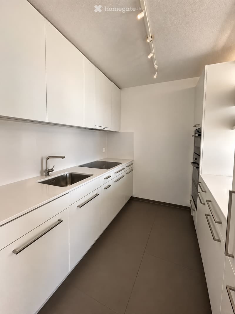 Modern, minimalist kitchen with white cabinets, stainless steel sink, and oven. Features sleek countertops and a dark tiled floor.