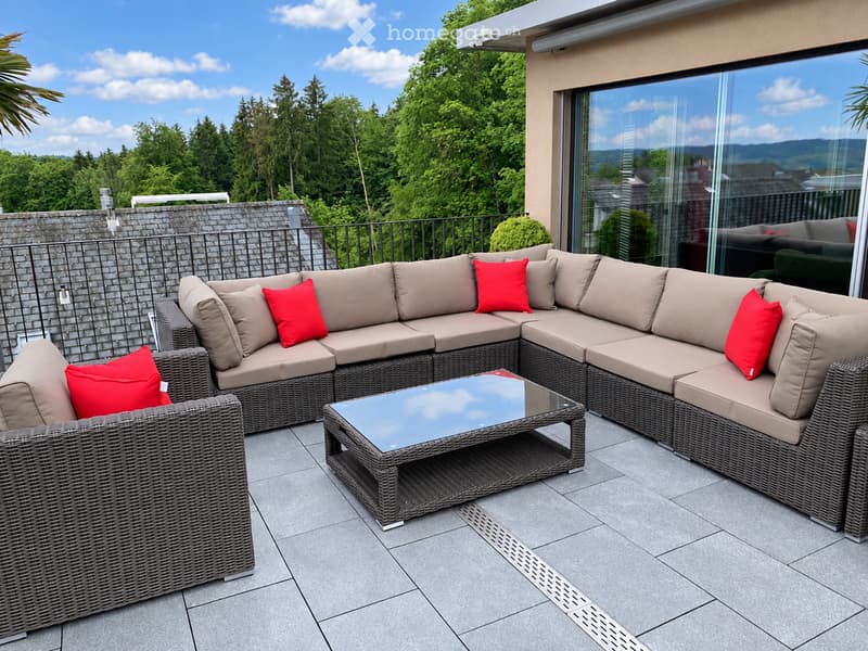 Outdoor patio with a modern wicker sectional sofa, red throw pillows, and a glass coffee table, overlooking a lush green landscape.