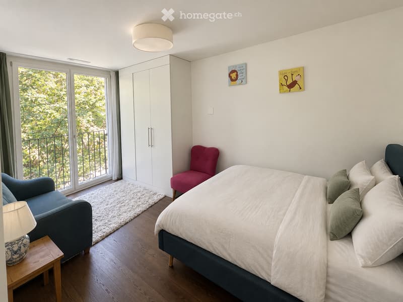 Cozy bedroom with a double bed, two armchairs, a small table with a lamp, a white cabinet, and a large window offering a view of greenery.