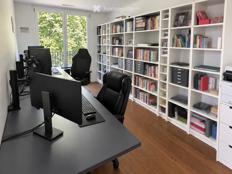 Modern office with a large desk, ergonomic chairs, dual monitors, keyboard, mouse, and a bookshelf filled with books and decorative items.