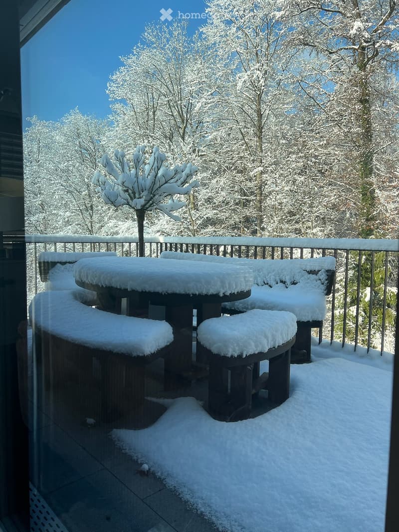 Snow-covered outdoor seating area with a table and chairs, surrounded by snow-laden trees and a clear blue sky.