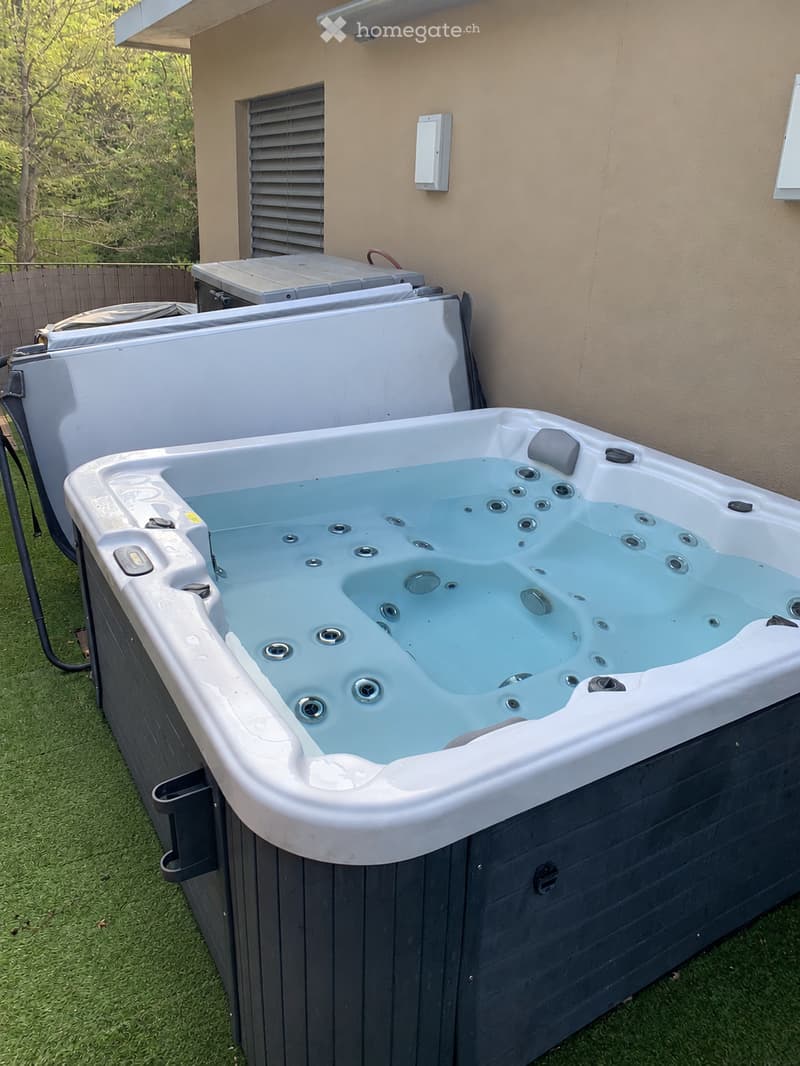 A modern hot tub with multiple jets and a control panel, situated on a grassy area next to a house with beige walls and a covered electrical unit.