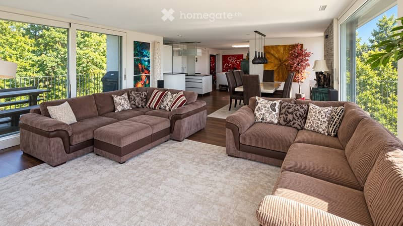 Modern living room with large windows offering a view of greenery, featuring a sectional sofa with pillows, an ottoman, a dining area with a table and chairs, and decorative art pieces.
