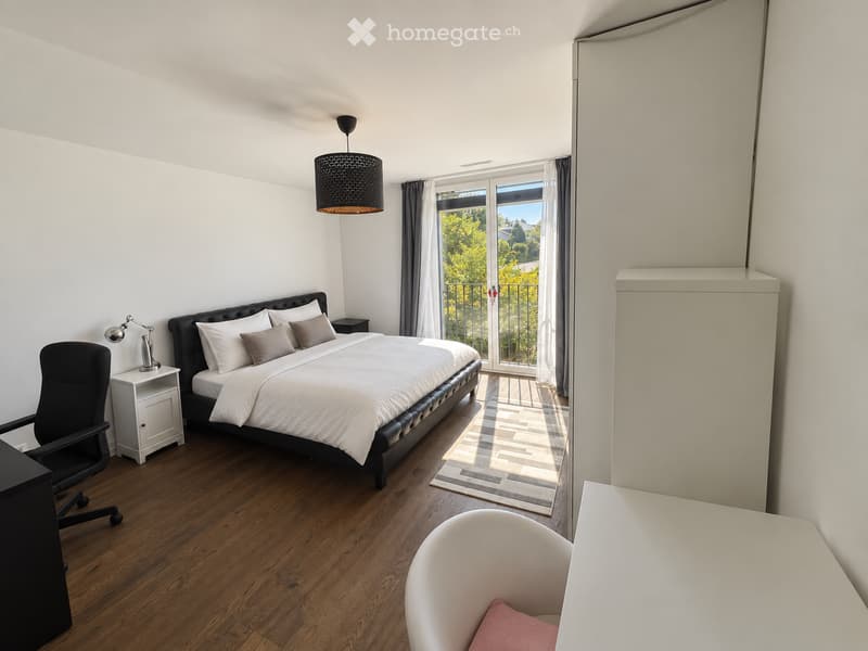 Modern bedroom with a large bed, nightstand, chair, and a view of greenery through large windows.