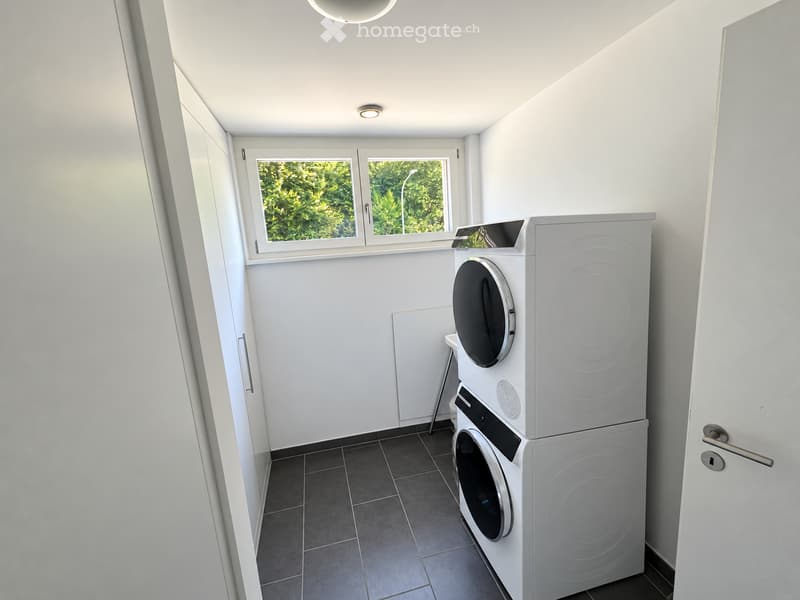 Modern bathroom with white walls, dark tiled floor, and a stacked washing machine and dryer unit near a window with a view of greenery.