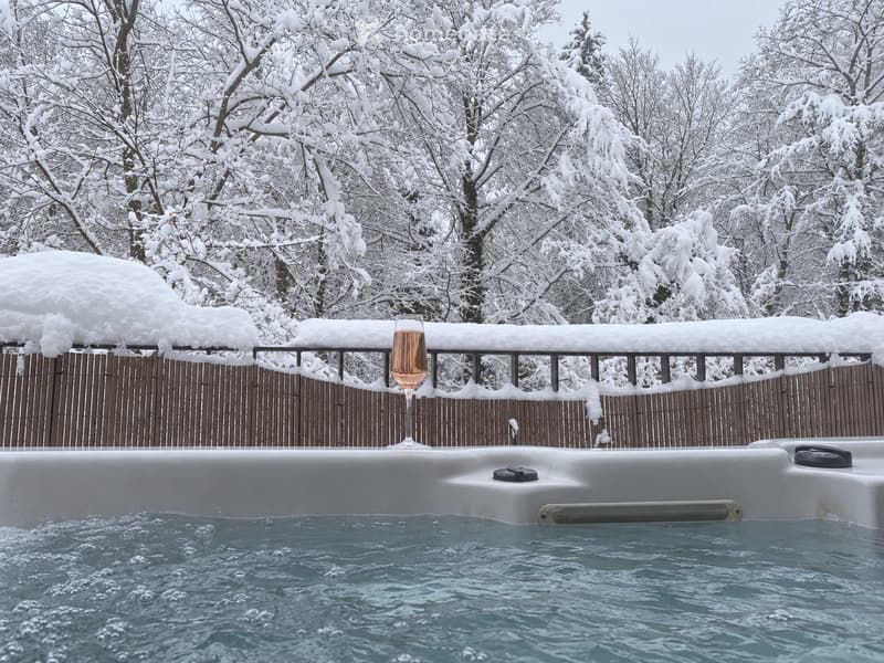 A hot tub surrounded by a wooden fence, with snow-covered trees in the background and a glass of champagne on the edge of the tub.