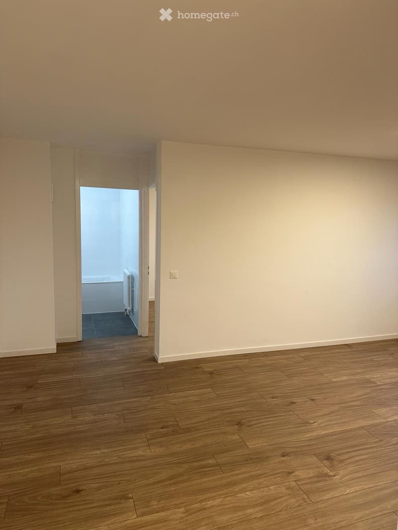 Empty room with wooden flooring and white walls, featuring an open doorway leading to another room with a bathtub visible in the background.