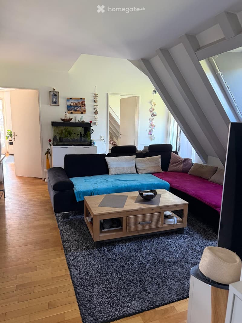 A modern living room with a sectional sofa, coffee table, fish tank, and cat litter. The room features wooden flooring, a large window, and a slanted ceiling typical of an attic space.