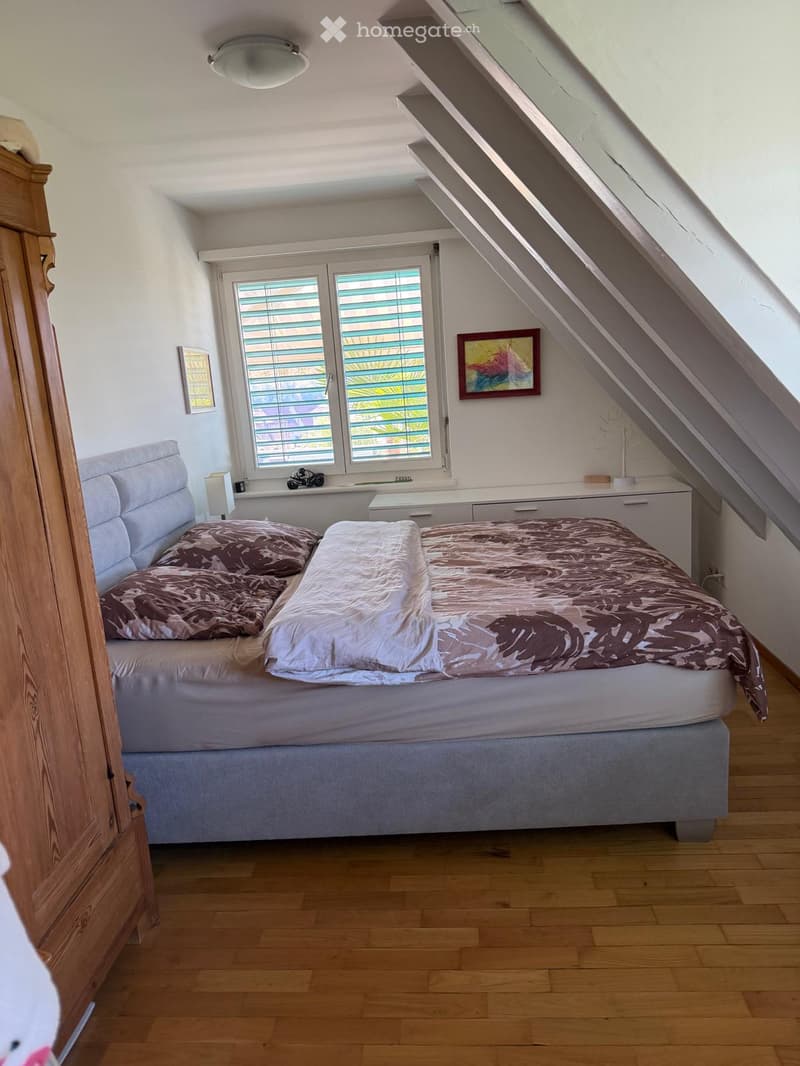 Cozy attic bedroom with wooden flooring, large window with blinds, bed with patterned bedding, nightstand with lamp, and a small decorative picture frame on the wall.