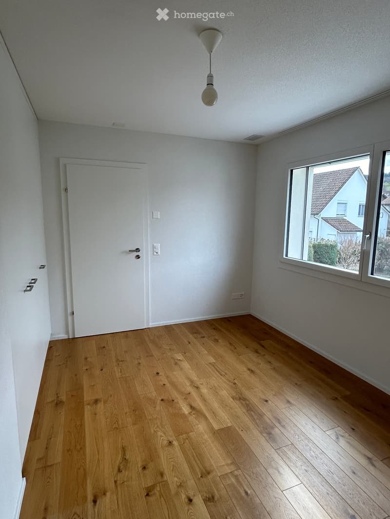 Empty room with light wooden flooring, white walls, a single window with a view of a neighboring house, and a white door.