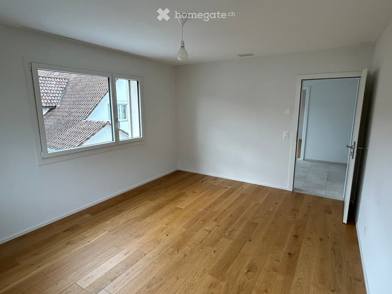 Empty room with wooden flooring, white walls, a large window showing a roof view, and an open doorway leading to another room.