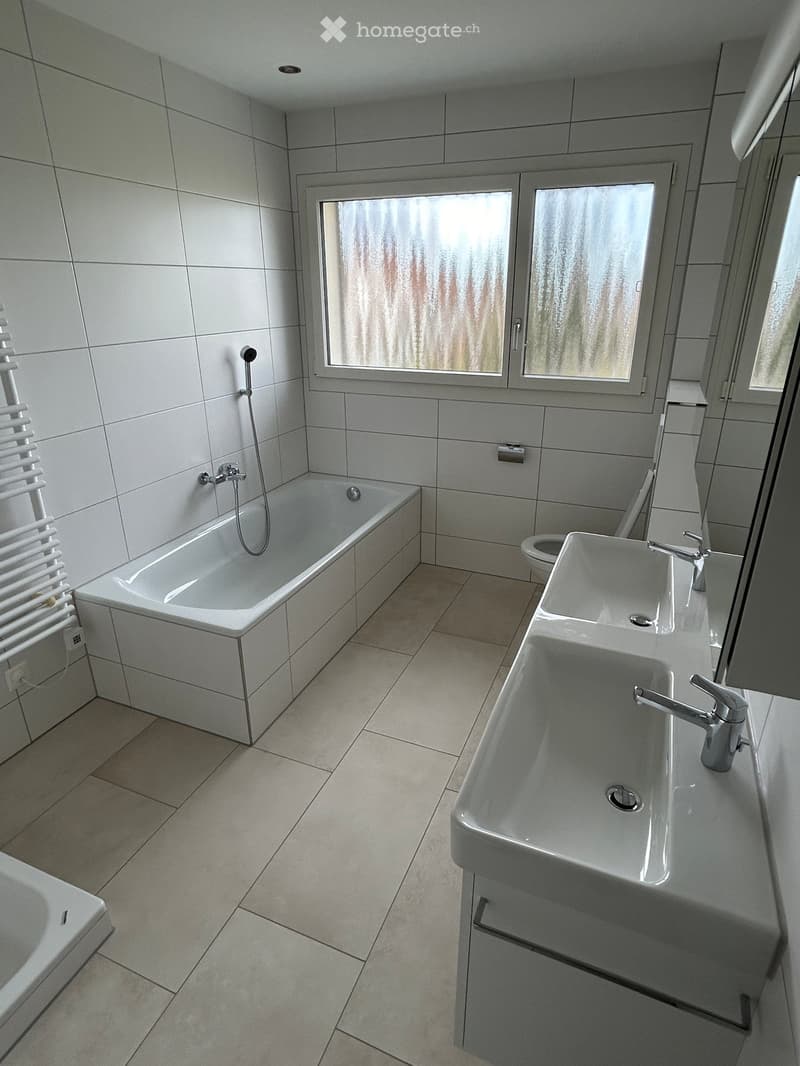Modern bathroom with white tiles, a bathtub, a sink with a faucet, a toilet, and a radiator. The room has large windows with frosted glass.