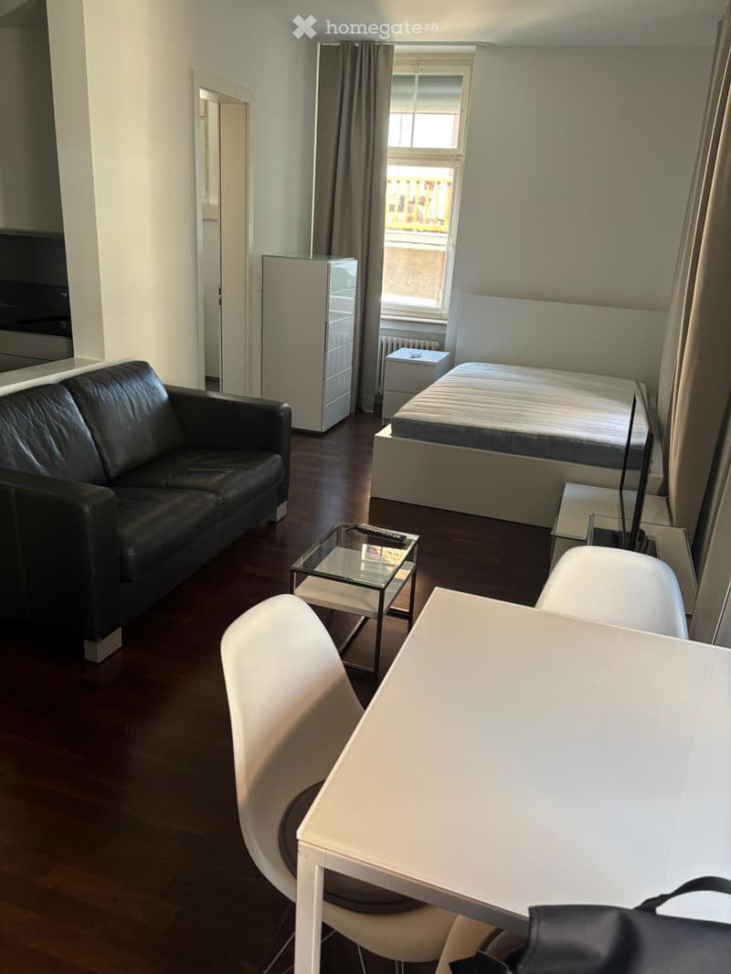 A modern, well-furnished room with a sofa, a bed, a small table, and a refrigerator. The room features wooden flooring and large windows with curtains.