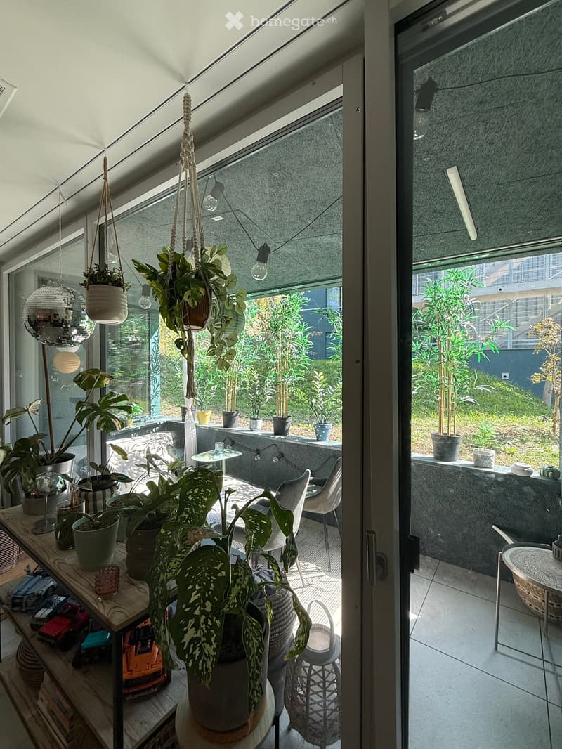 A modern, well-lit room with large windows and a glass door leading to a lush garden. The space features a wooden shelf with potted plants, a small round table, and several hanging plants. The room is decorated with a mirror and a few chairs, creating a c
