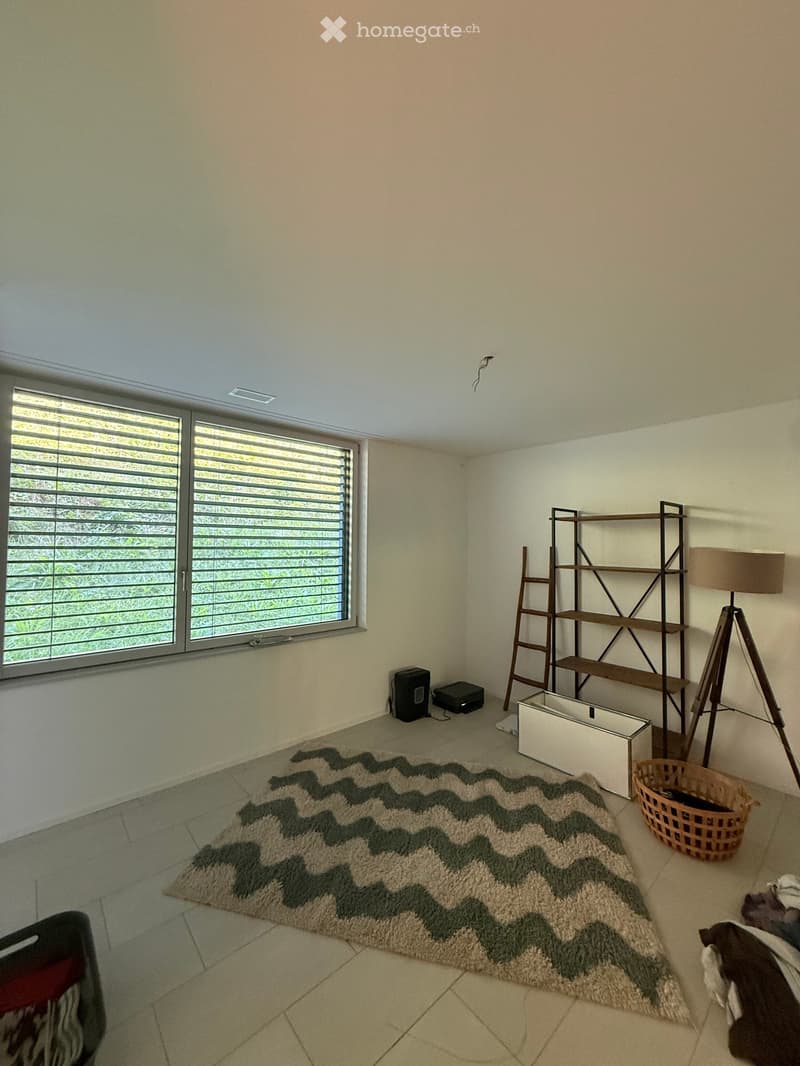 Empty room with tiled floor, large window with blinds, shelf, floor lamp, rug, and a small basket.