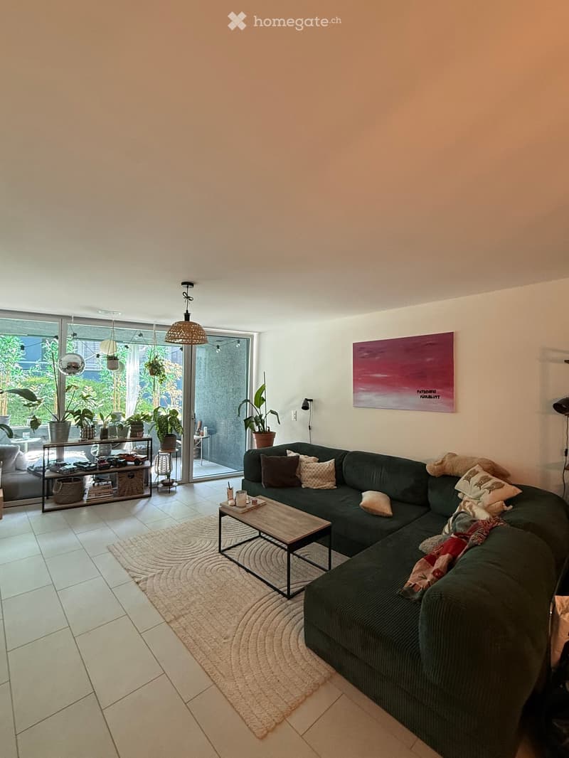 Modern living room with a green sectional sofa, wooden coffee table, area rug, shelf with potted plants, and abstract art on the wall.
