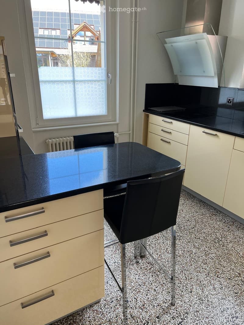 Modern kitchen with light-colored cabinets, black countertops, a black dining table with chairs, and a stove with a hood.