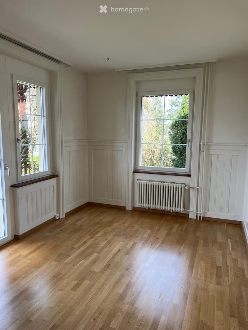 Empty room with light wooden flooring, white walls, two windows with white frames, and a radiator under one window.
