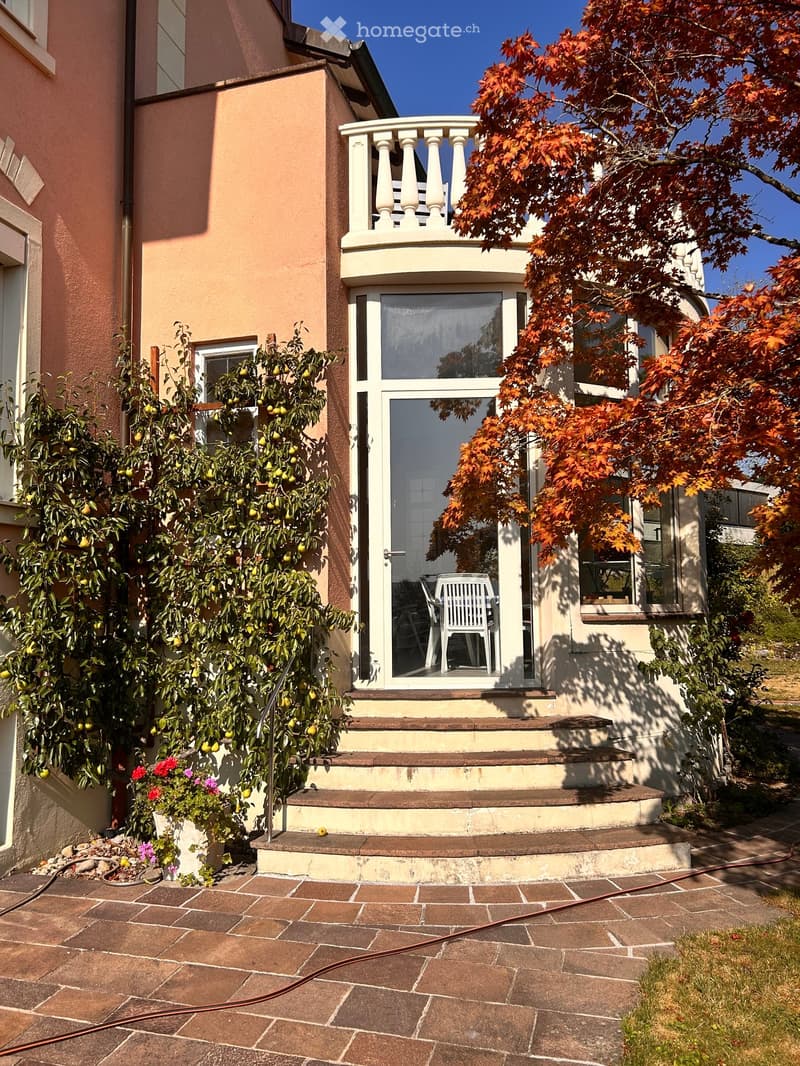 Two-story house with a raised entrance, balcony, and surrounding greenery including fruit trees and flowering plants.
