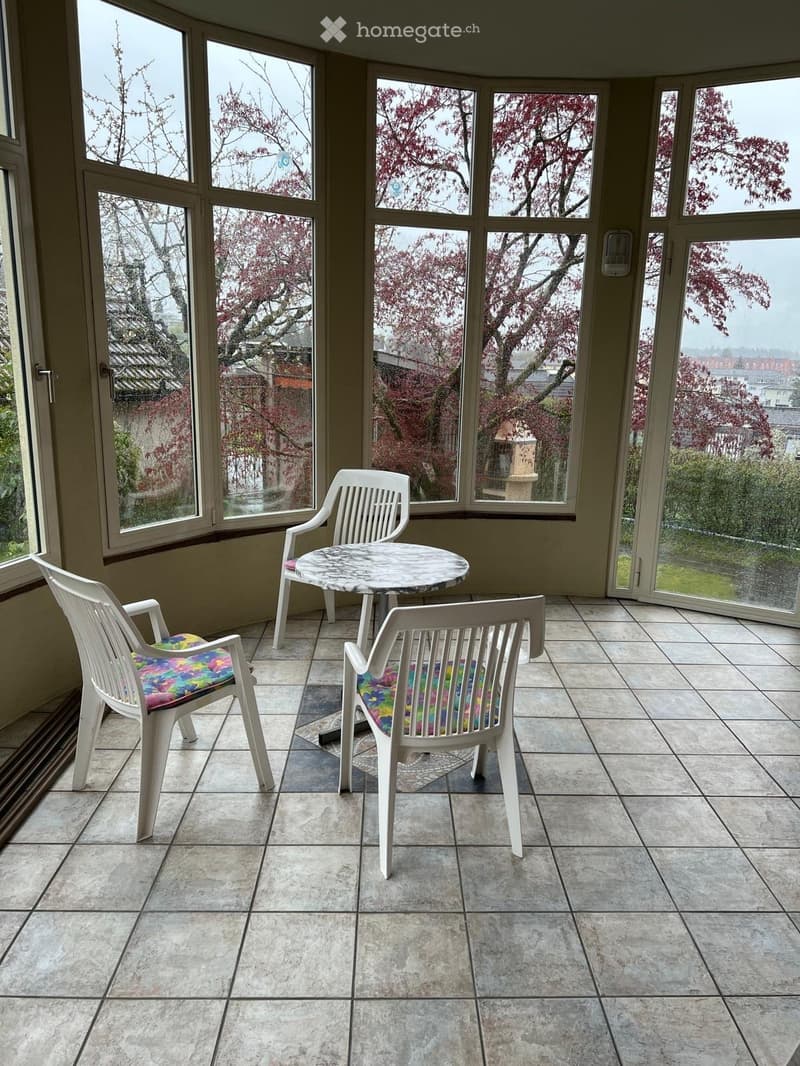 Brightly lit balcony with large windows, tiled floor, and a small round table with four colorful chairs.
