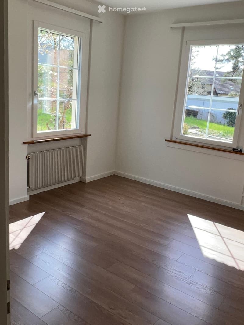 Empty room with wooden flooring, white walls, and large windows with wooden sills. Radiator visible under one window.