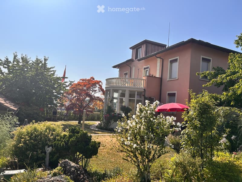 Two-story pink house with white trim, balcony, and surrounding garden with trees and flowers.
