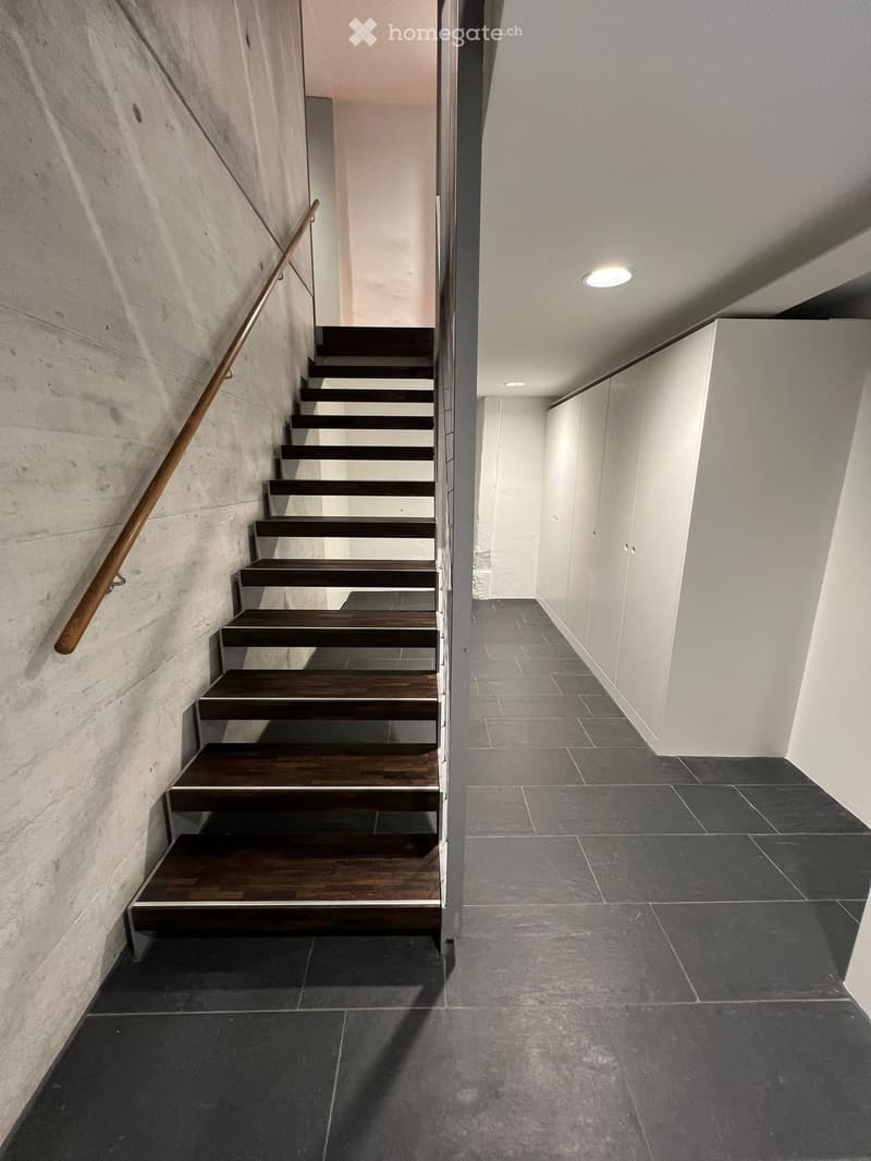 A modern staircase with wooden steps and a metallic handrail, on the left side, an interior space with a white wall, and a tiled floor.