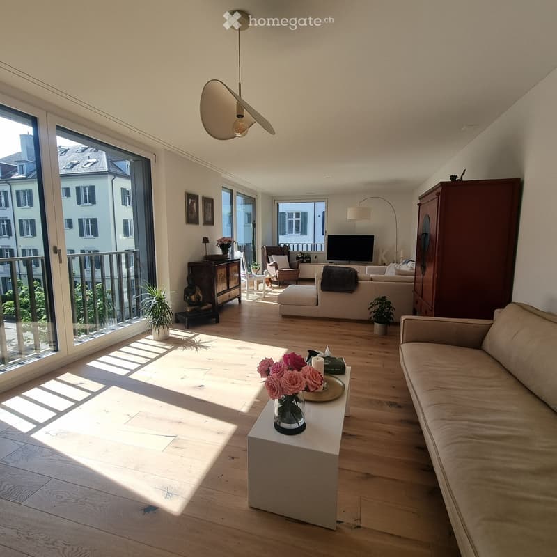 Bright living room with wooden flooring, large windows, and a balcony. Features a sofa, coffee table, armchair, ottoman, floor lamp, table lamp, television, cabinet, vase with flowers, and plants.