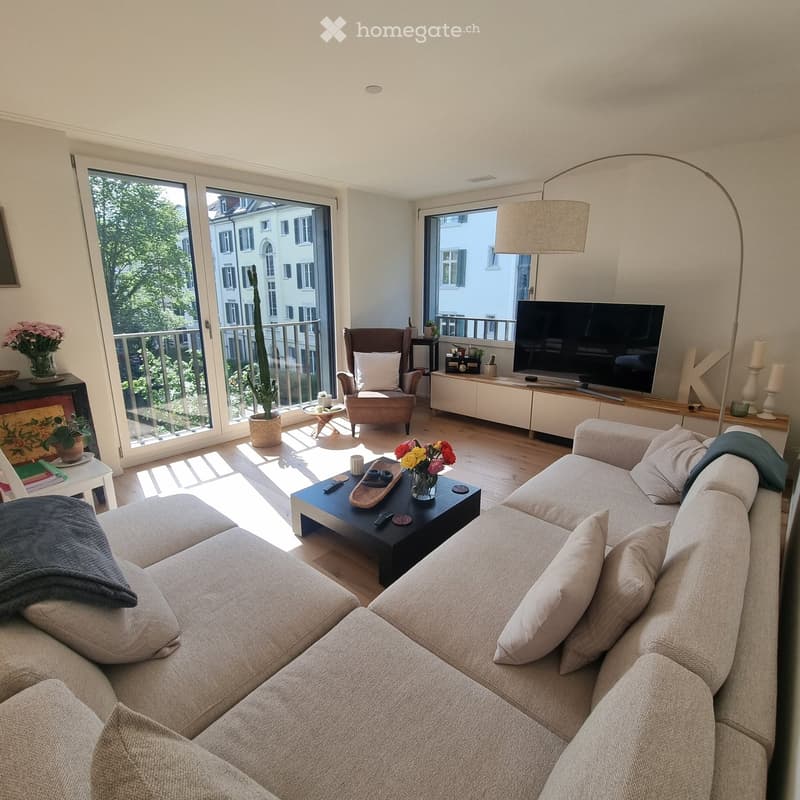 Modern living room with large windows and balcony access, featuring a grey sectional sofa, coffee table, armchair, TV stand, floor lamp, and decorative items.