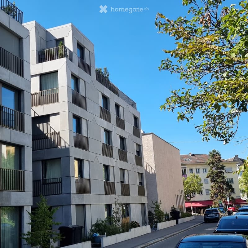 Modern multi-story apartment building with balconies, large windows, and contemporary design, situated on a street with trees and parked cars.
