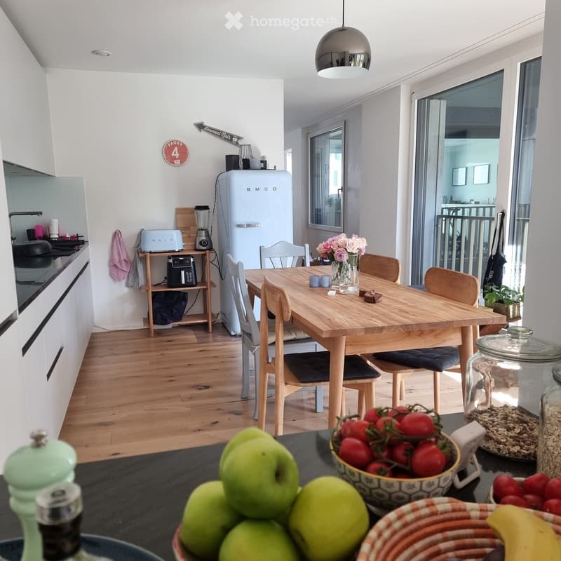 Bright, modern kitchen with wooden flooring, white cabinets, and a central dining table surrounded by chairs. The kitchen features a refrigerator, blender, toaster, and various decorative items including a vase with flowers and bowls of fruit on the count