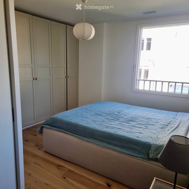A well-lit bedroom with a single bed, a wardrobe, and a hanging lamp. The room has wooden flooring and a large window with a view.