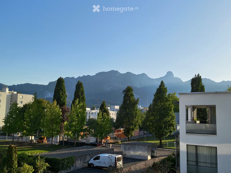 Modern building with a balcony overlooking a landscaped area, surrounded by tall trees and distant mountains.