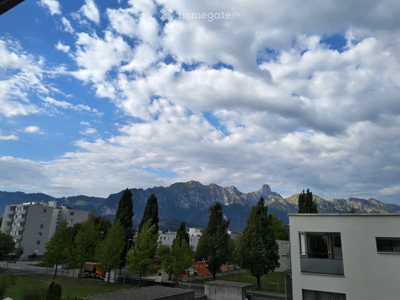 View of a residential area with modern buildings, tall trees, and a backdrop of mountain peaks under a partly cloudy sky.