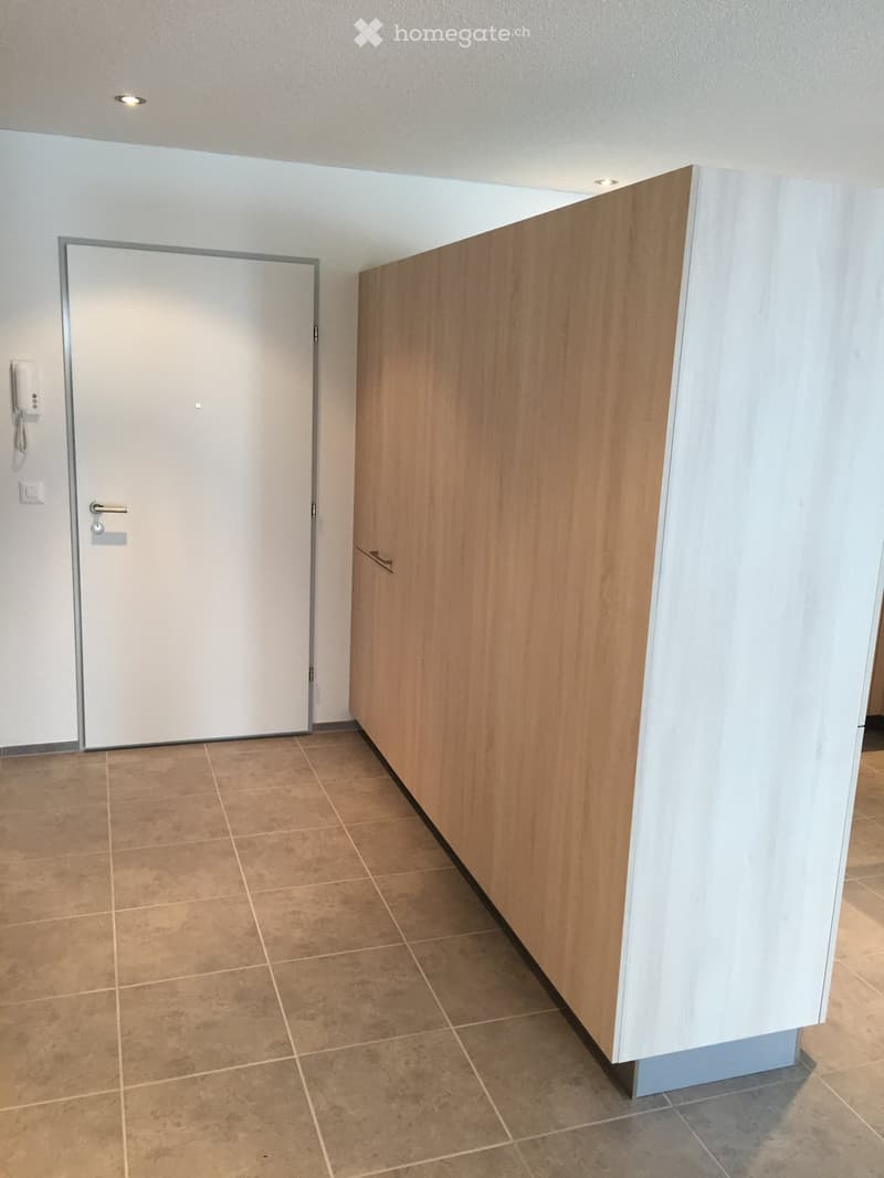 Empty hallway with tiled floor, white walls, and a wooden cabinet on the right side. A closed door is visible on the left.