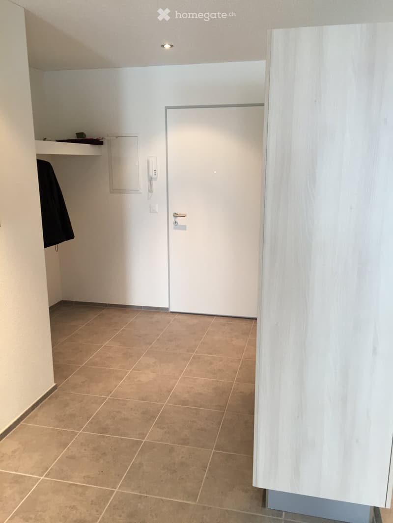 Empty hallway with tiled floor, white walls, and a closed door. A coat is hanging on the wall to the left.