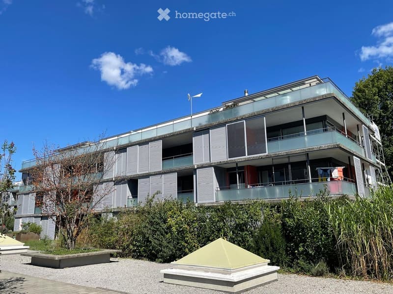 Modern multi-story building with glass balconies and white shutters, surrounded by greenery and a small garden area with a pyramid-shaped structure.