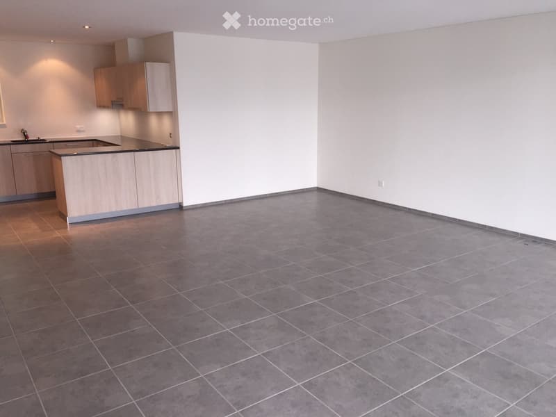 Empty room with grey tiled floor and white walls, featuring a small kitchen area with cabinets and a sink.