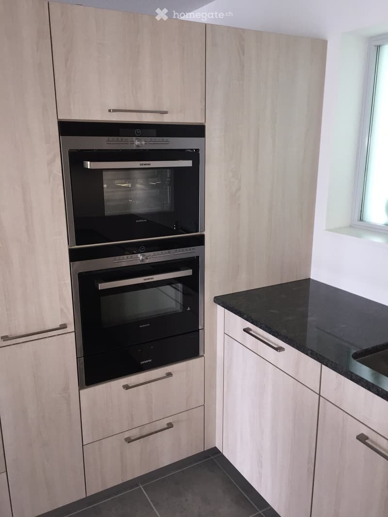 Modern kitchen with light wood cabinets, black countertop, and built-in Siemens ovens.