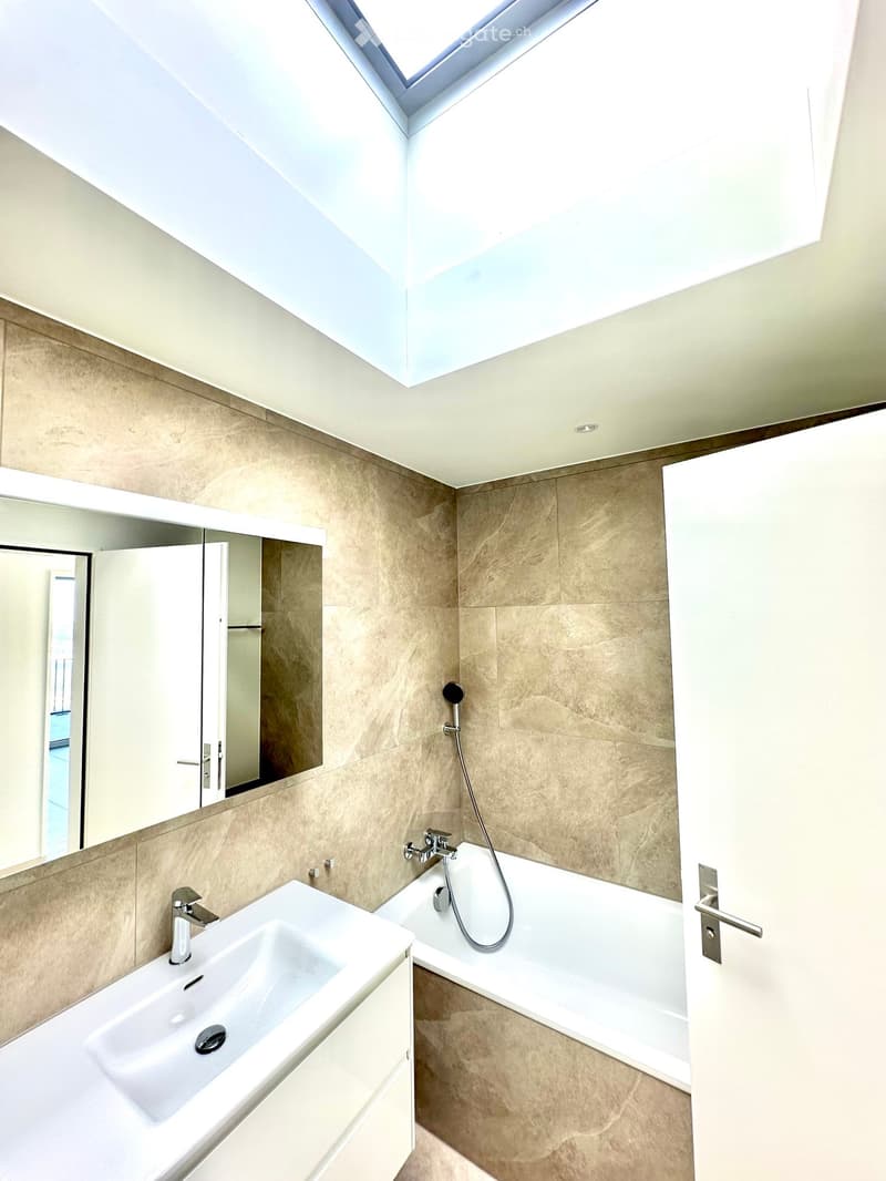 Modern bathroom with a large skylight, beige tiled walls, a white sink, a bathtub with a handheld shower, and a large mirror with built-in storage.