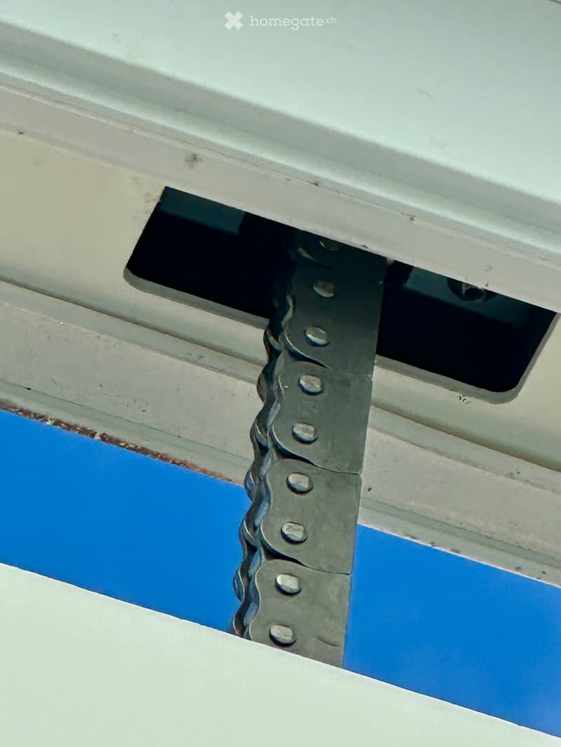 Close-up of a metal hinge mechanism attached to a white surface, likely part of a boat or trailer structure, with visible bolts and a blue painted section below.