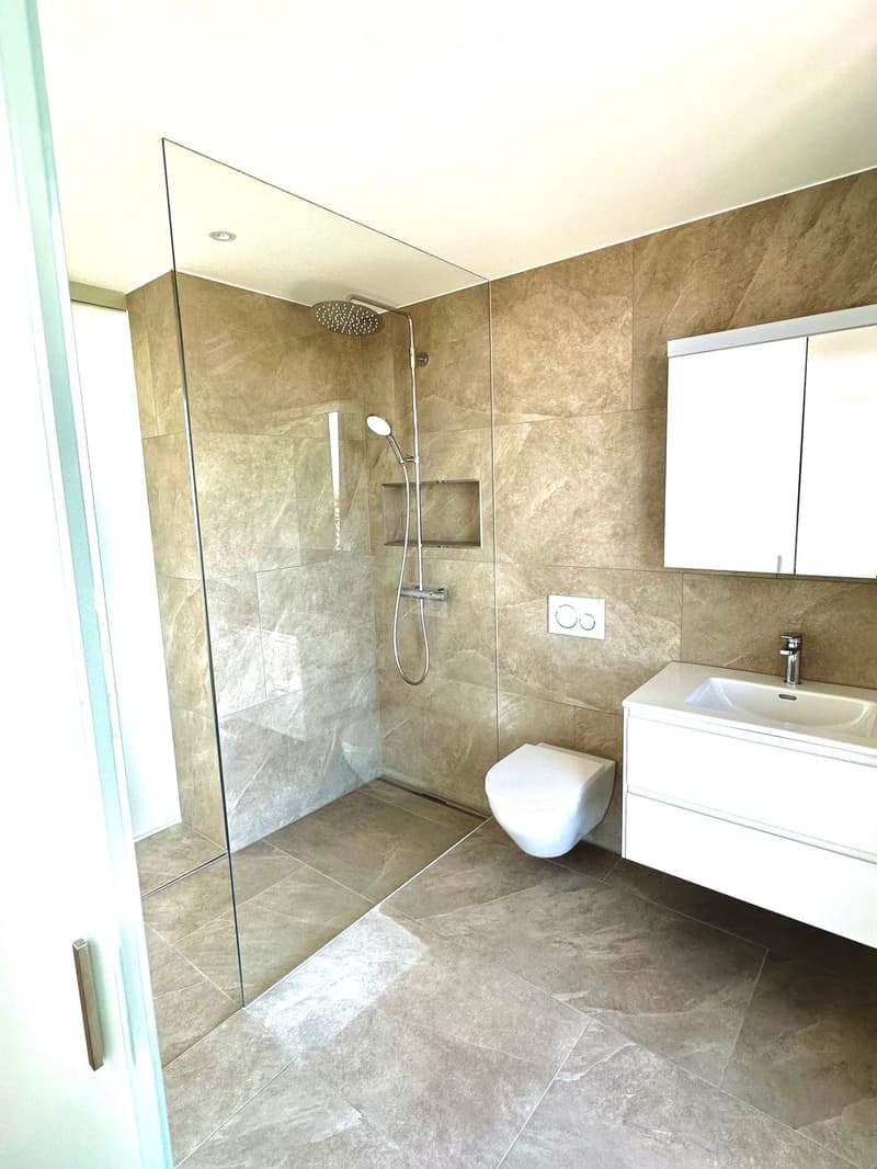 Modern bathroom with beige tiled walls and floor, glass-enclosed shower, toilet, sink with cabinet, and mirror.