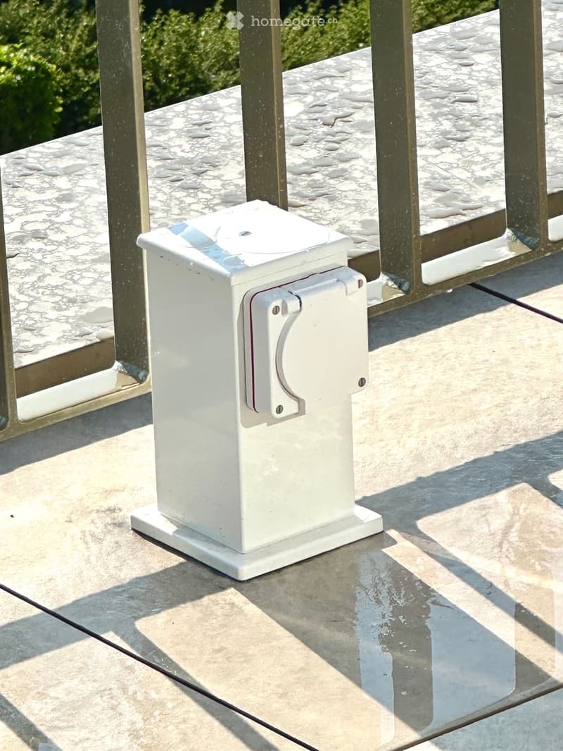 A white electrical outlet mounted on a marble balcony railing, with a metal frame and a wet surface indicating recent rain.