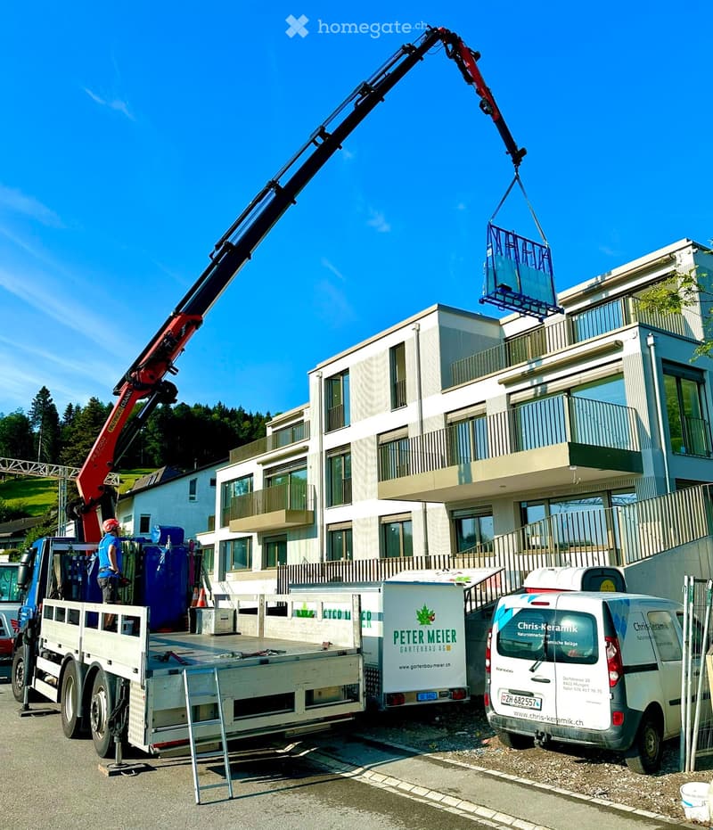 A modern multi-story building with large windows and balconies, with a crane lifting a container onto the roof.
