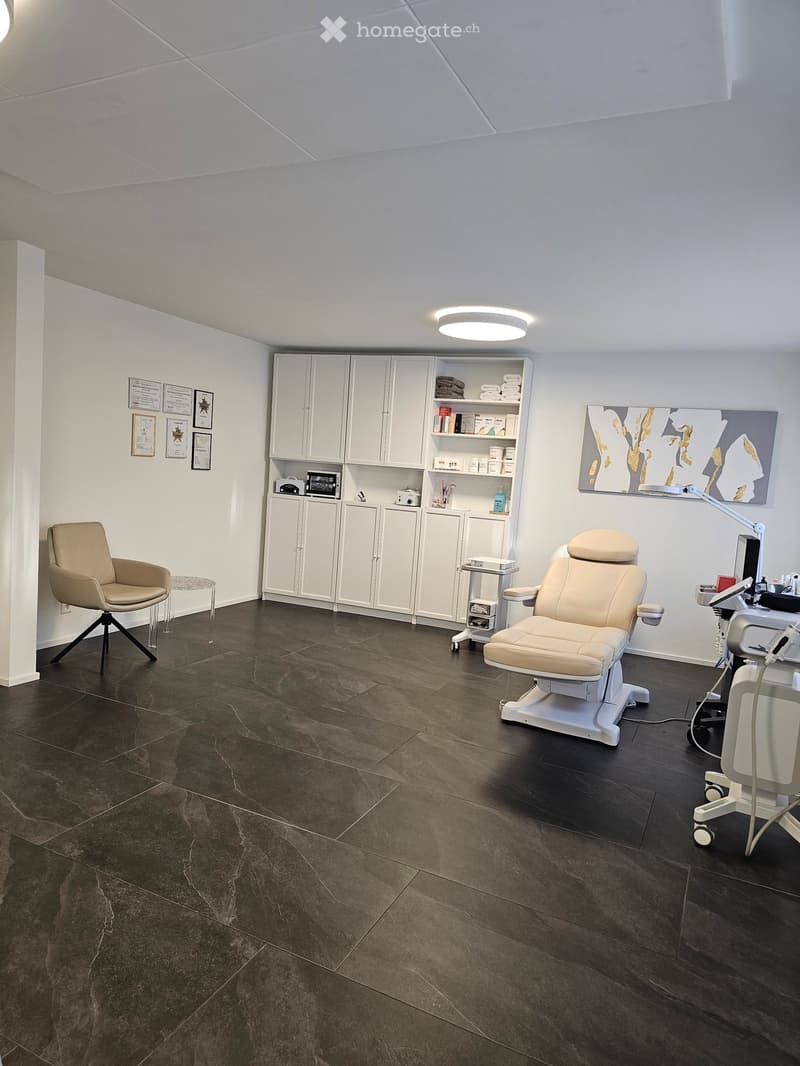 A modern, well-lit room with a dark tiled floor, white cabinets, a beige chair, a small stool, a mirror, a lamp, and a screen.